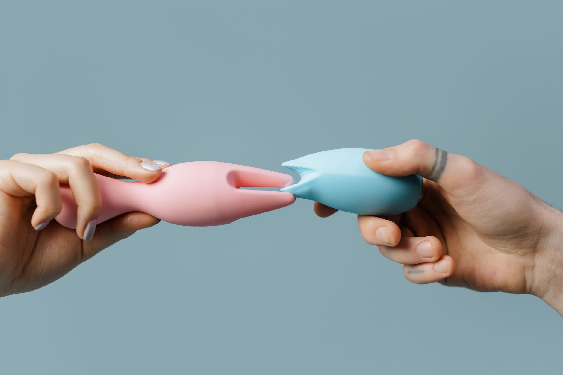 Two hands holding pink and blue silicone vibrators against a pastel background.