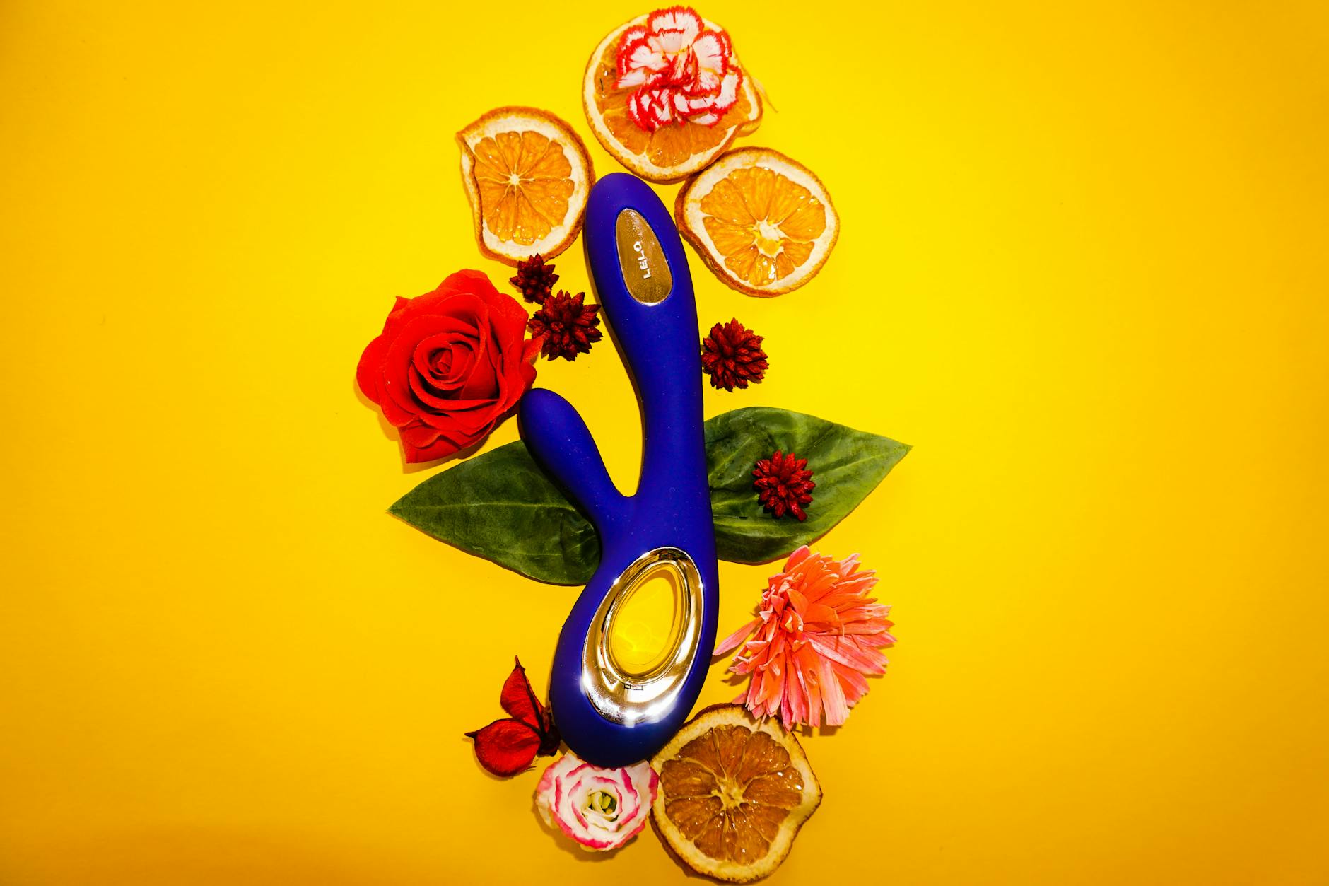 A blue lemon vibrator surrounded by flowers and citrus slices on a bright yellow background, symbolizing fresh approaches to pleasure.